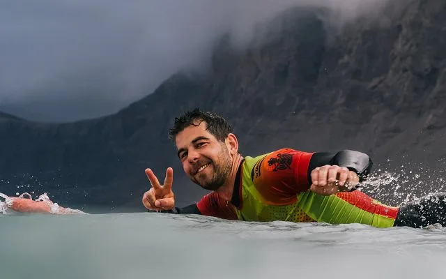 Uśmiechnięty surfer wiosłuje w Famarze, w tle klify Risco