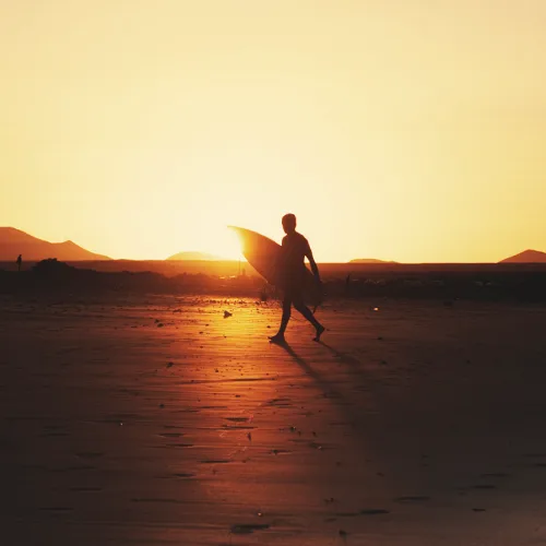 Surfer z deską o zachodzie słońca na plaży Famara, surf camp na Lanzarote